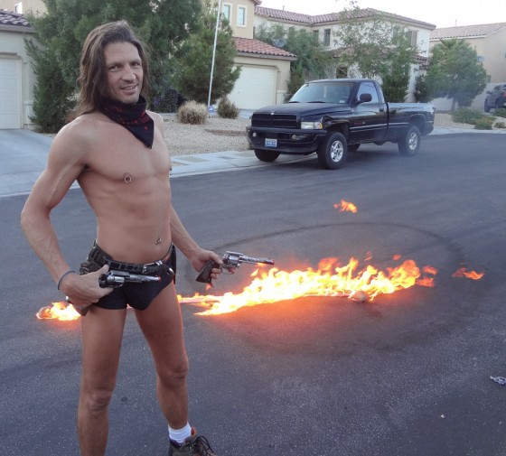 Shirtless and tan male burlesque dancer Kevin Matthews with pistol and fire on asphalt behind him