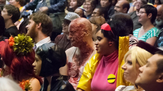 Horribly burnt Zombie cosplayer Andrew Myers next to smiling yellow trenchcoat wearing Jubilee in crowd at costume contest
