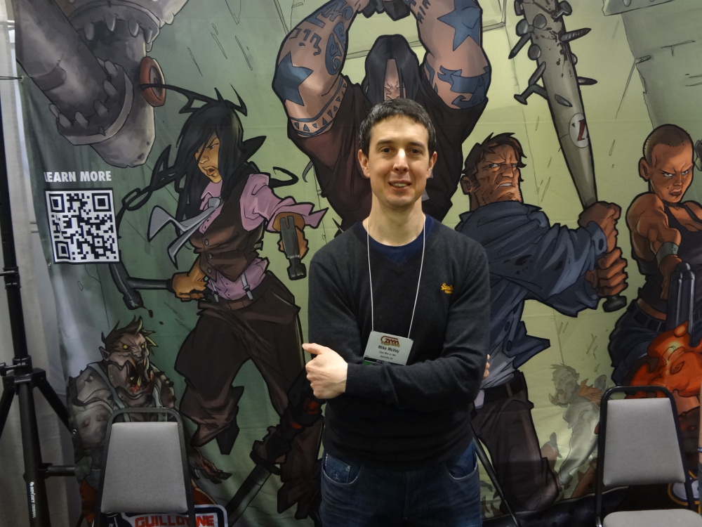 Thin British painter Mike McVey with crossed arms in front of Zombicide poster at Cool Mini booth
